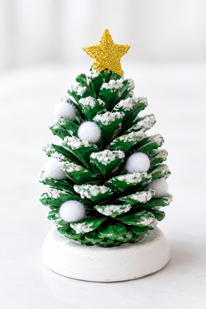 A green-painted pinecone Christmas tree with white pom-poms and a gold star on a white surface.
