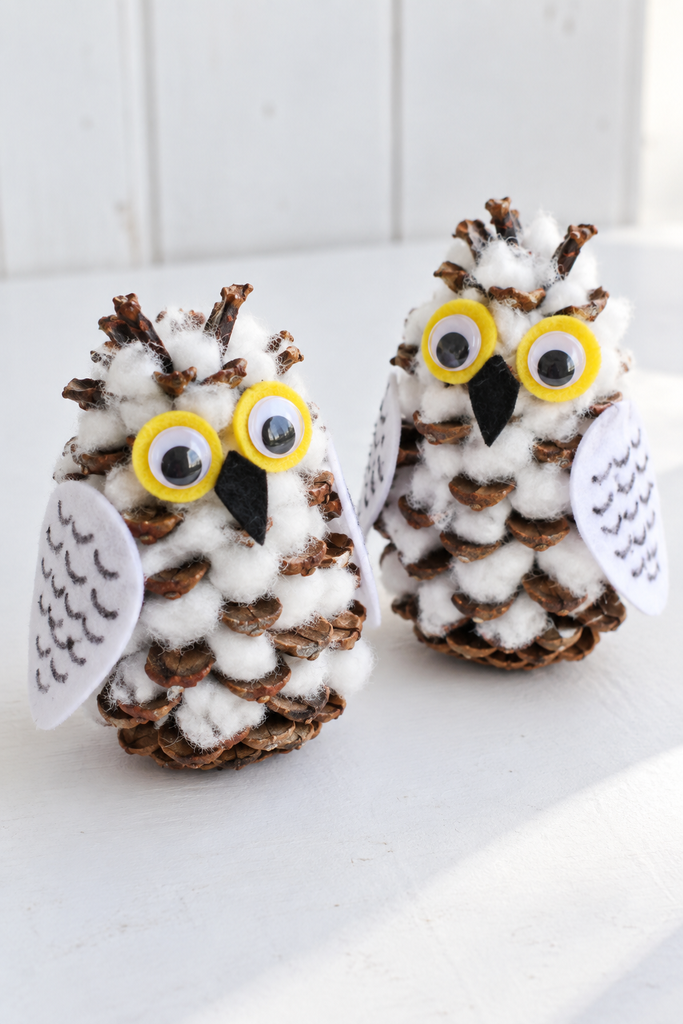 Two cotton-stuffed pinecone owls with yellow felt eyes and white felt wings standing on a white table.