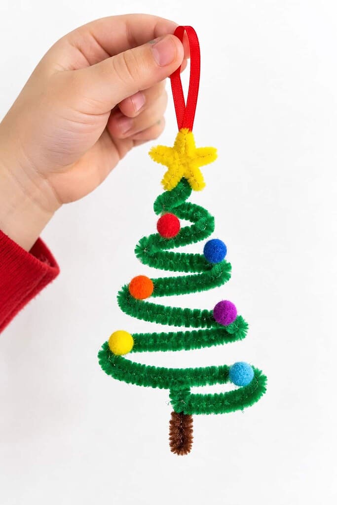 A child's hand holding a green zigzag pipe cleaner tree ornament with pom-poms and a yellow star.