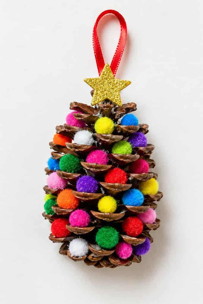 a pinecone ornament craft with colorful mini pom-poms tucked between the scales, a small topper, and a ribbon hanging loop on a white table
