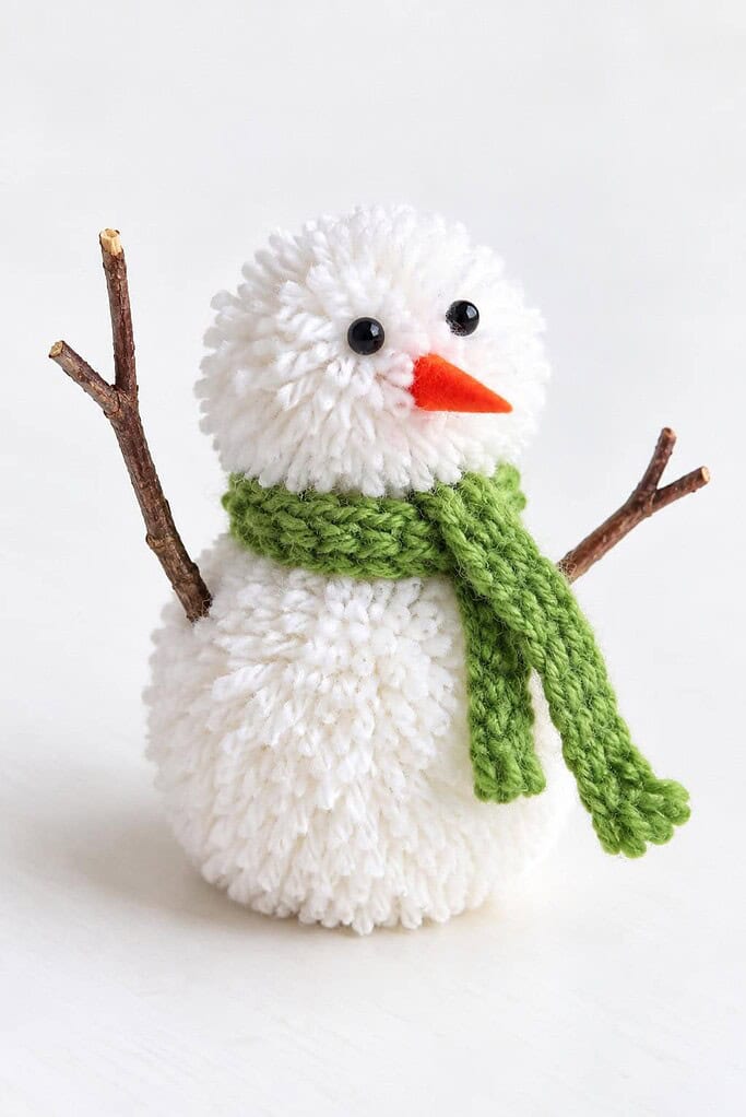 a fluffy white yarn pom-pom snowman with a knit scarf, bead eyes, an orange nose, and twig arms on a white table