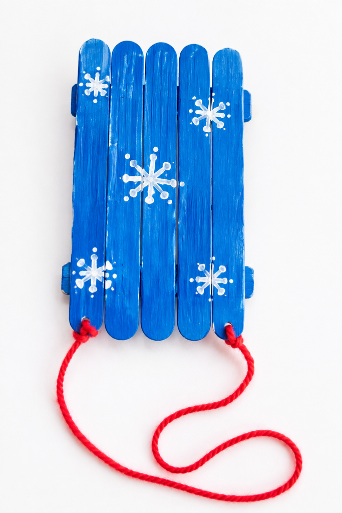 a sled craft made from craft sticks painted bright colors with white snowflakes and a yarn pull rope on a white table