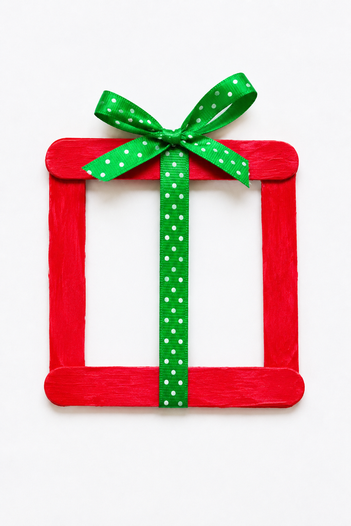 a Christmas present craft made from craft sticks glued into a painted square frame with a ribbon up the middle and a bow on top on a white table