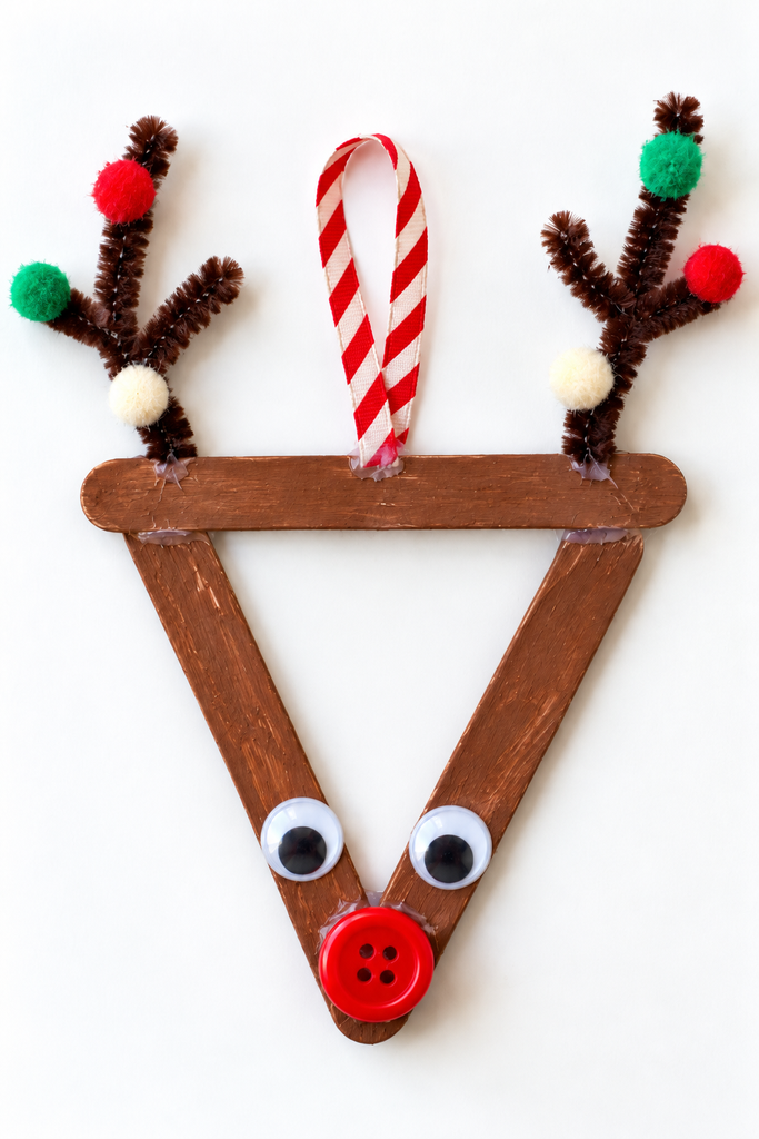 a craft stick reindeer ornament in a downward triangle with pipe cleaner antlers, colorful pom-poms, googly eyes, and a red button nose on a white table