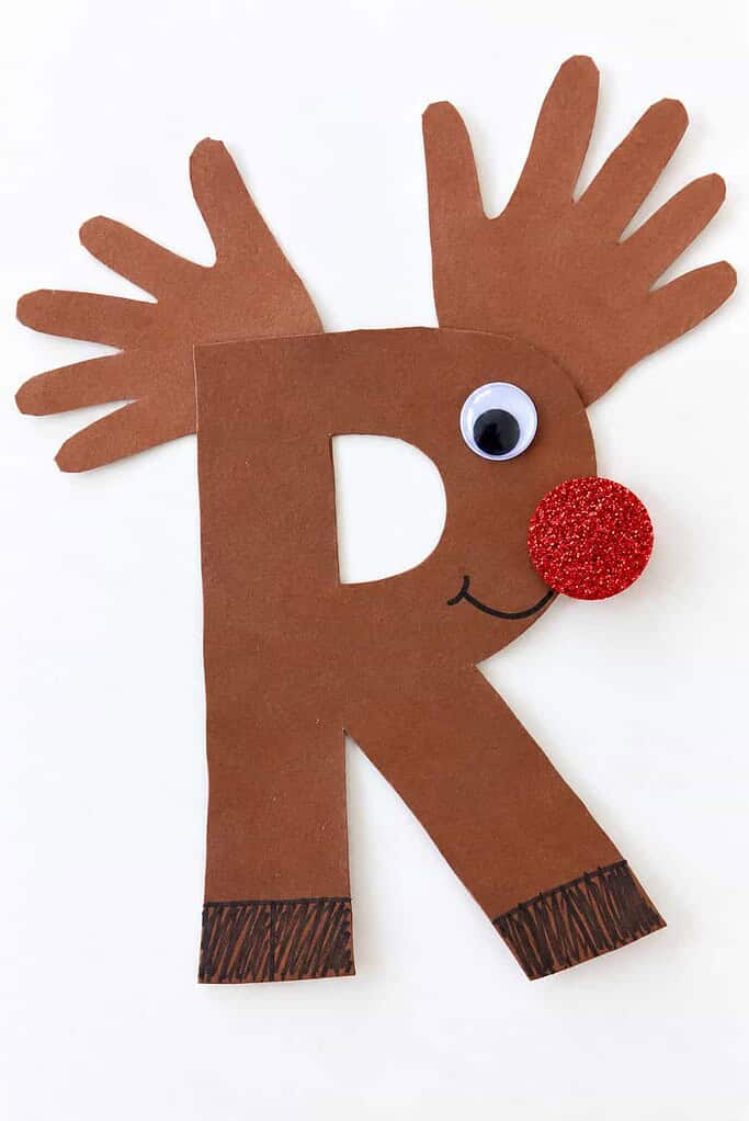 a brown paper letter R reindeer craft with handprint antlers, a googly eye, a smile, and a red glitter nose on a white table