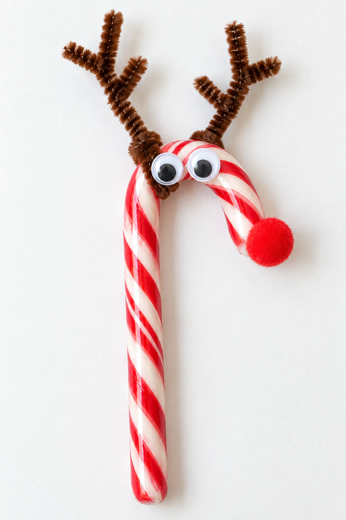 a candy cane reindeer craft with brown pipe cleaner antlers, googly eyes, and a red pom-pom nose on a white table