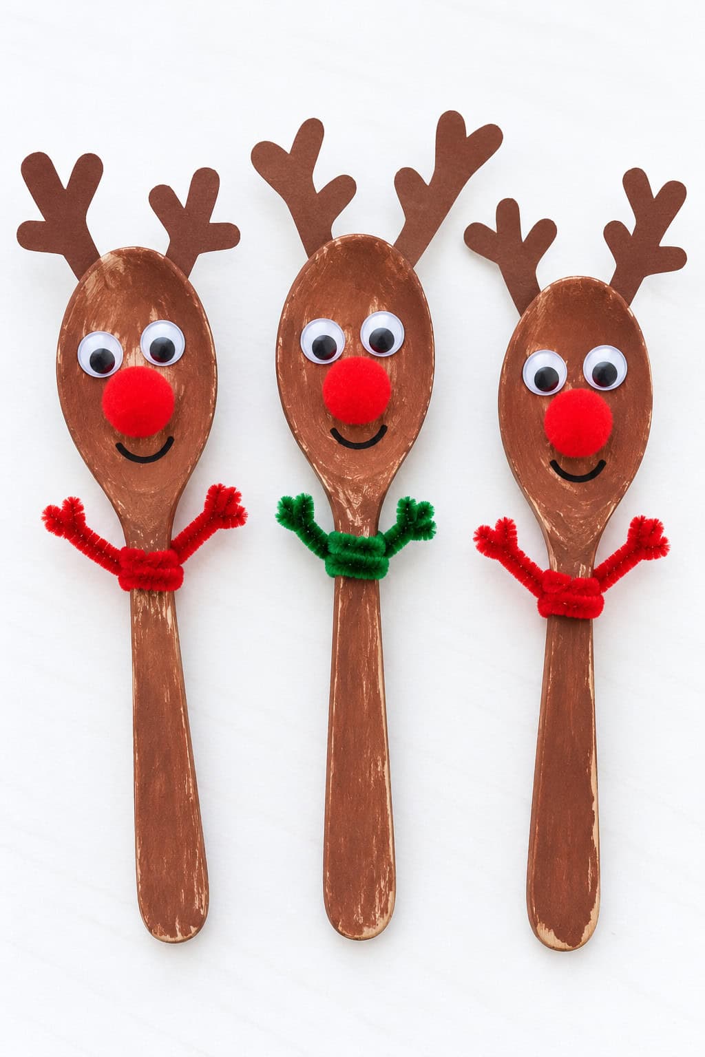 three handmade wooden spoon reindeer crafts painted brown with antlers, googly eyes, and red pom-pom noses on a white table