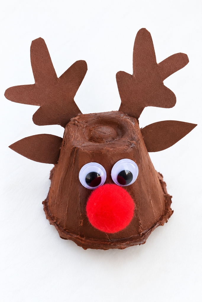a brown egg carton cup reindeer with paper antlers, googly eyes, and a red pom-pom nose on a white table