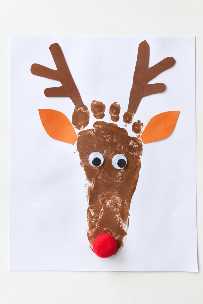 a reindeer footprint craft made from a brown painted footprint with paper antlers, orange ears, googly eyes, and a red pom-pom nose on a white table
