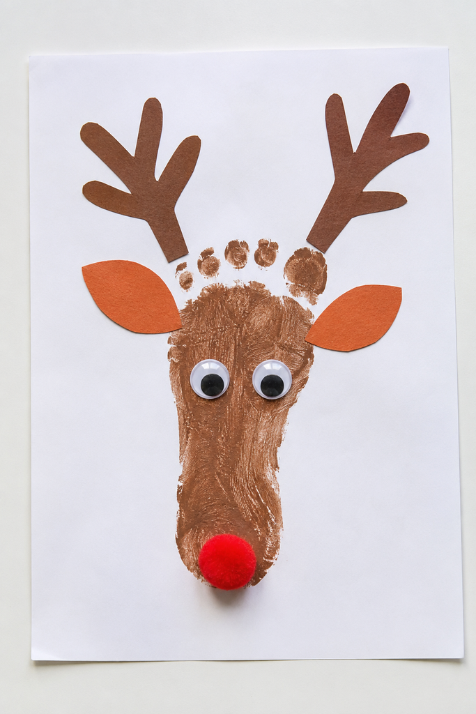 a reindeer footprint craft made from a brown painted footprint with paper antlers, orange ears, googly eyes, and a red pom-pom nose on a white table
