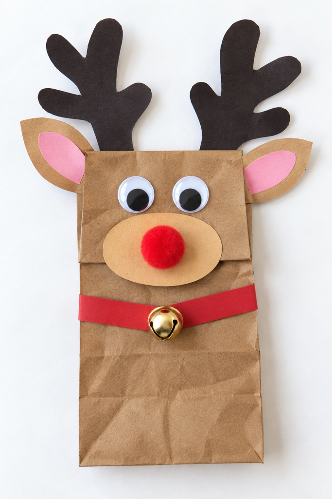 a brown paper bag reindeer puppet with black paper antlers, googly eyes, a red pom-pom nose, and a jingle bell collar on a white table