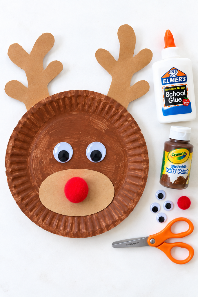 a brown paper plate reindeer craft with paper antlers, a muzzle, googly eyes, and a red pom-pom nose, with scissors, glue, and paint staged beside it on a white table