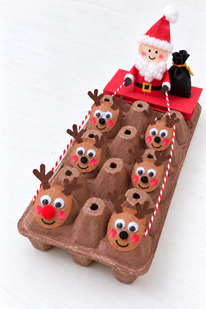 an egg carton reindeer sleigh craft with a flat red paper sleigh, a little Santa figure, and a team of round egg-cup reindeer with googly eyes and antlers joined by twine reins on a white table
