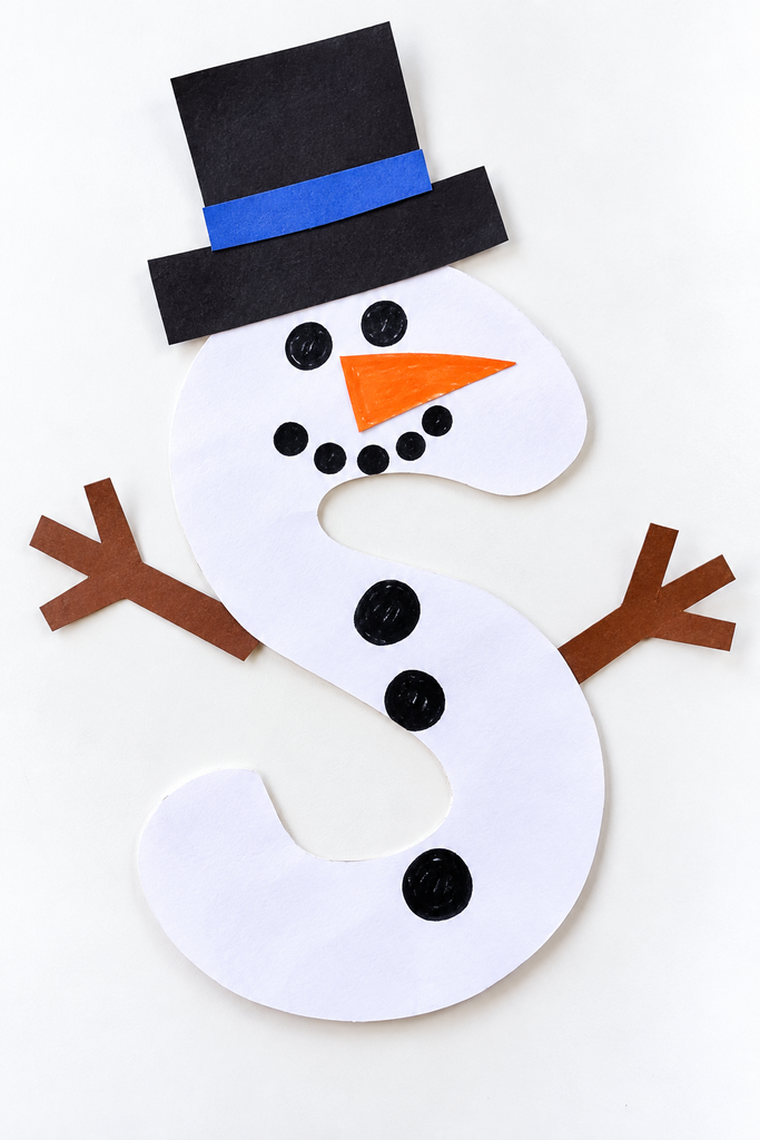 a white paper letter S snowman craft with a black hat, dot eyes, an orange nose, dot buttons, and twig arms on a white table