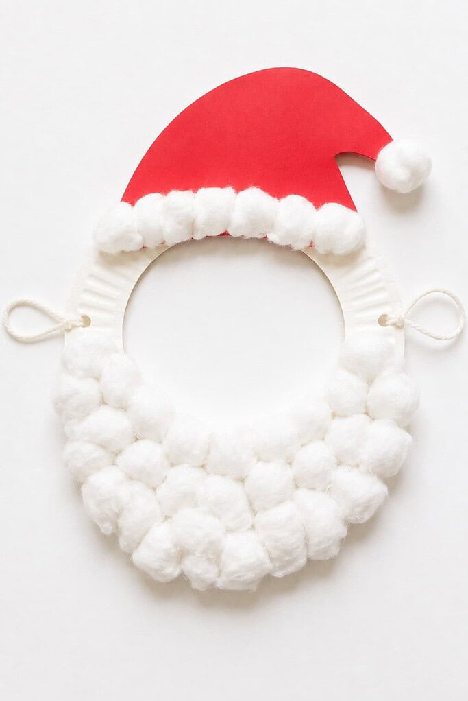 a wearable Santa beard craft made from a paper plate with a face hole, a fluffy cotton ball beard, a red paper hat, and string ties on a white table