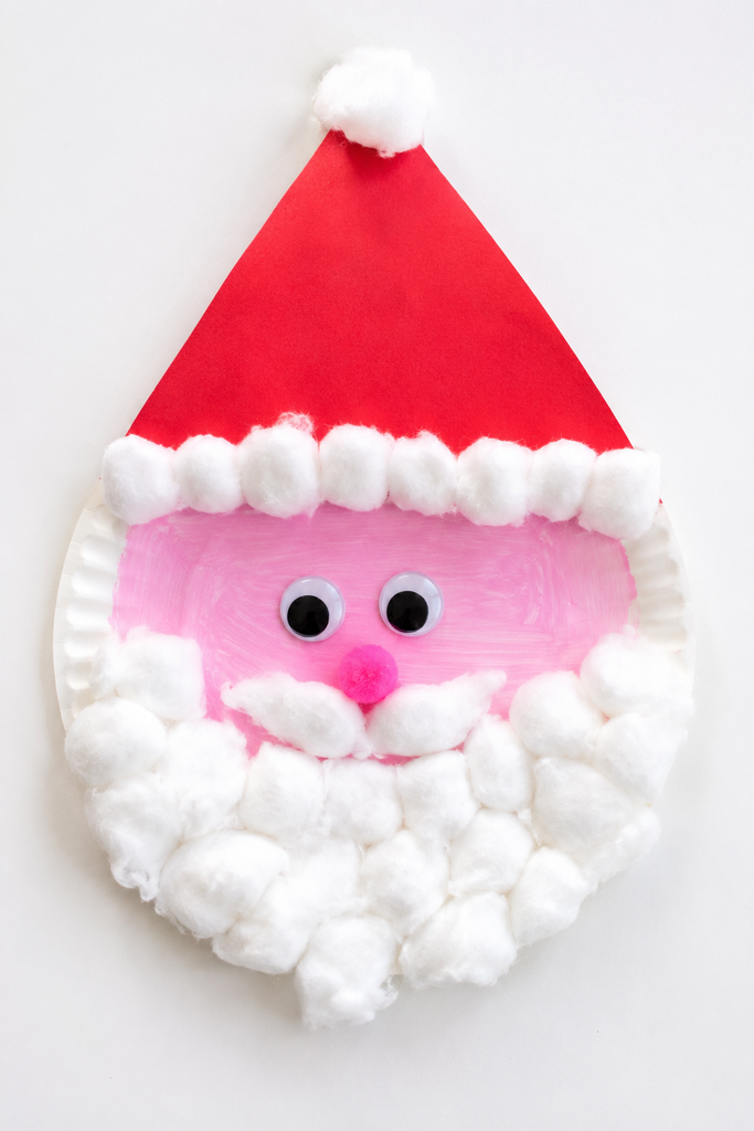 a paper plate Santa Claus craft with a red triangle hat, a cotton beard and mustache, googly eyes, and a pink pom-pom nose on a white table