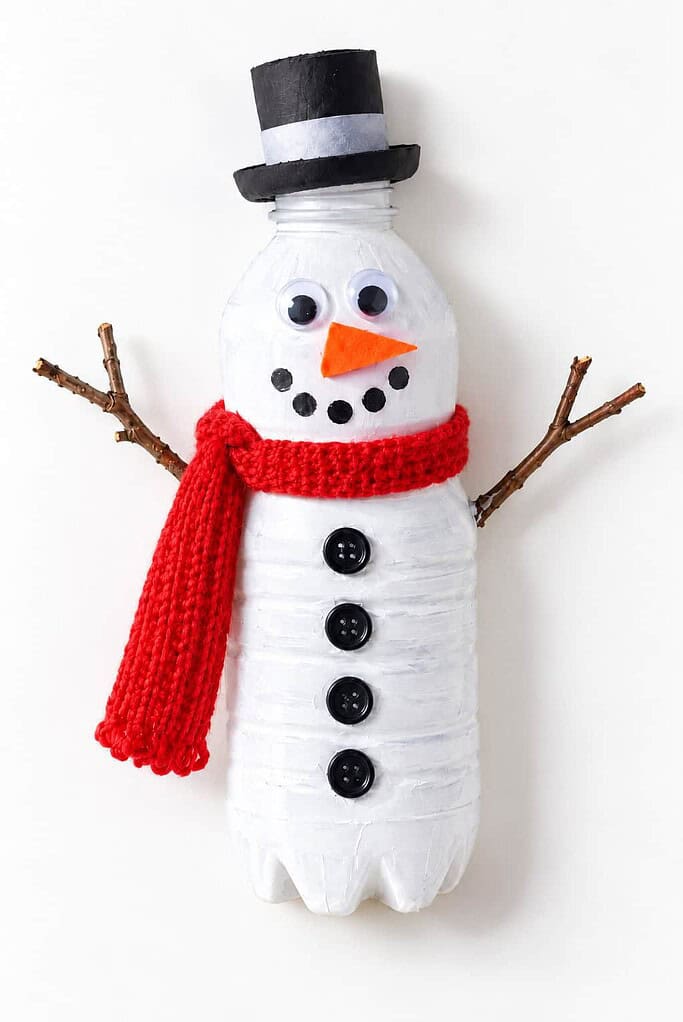 a white plastic bottle snowman with a black hat, googly eyes, an orange nose, buttons, a scarf, and twig arms on a white table
