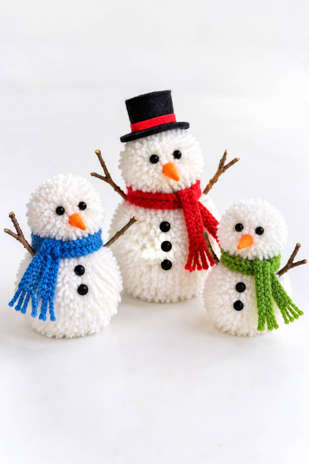 three fluffy white pom-pom snowman crafts with knit scarves, bead eyes, orange noses, and twig arms on a white table