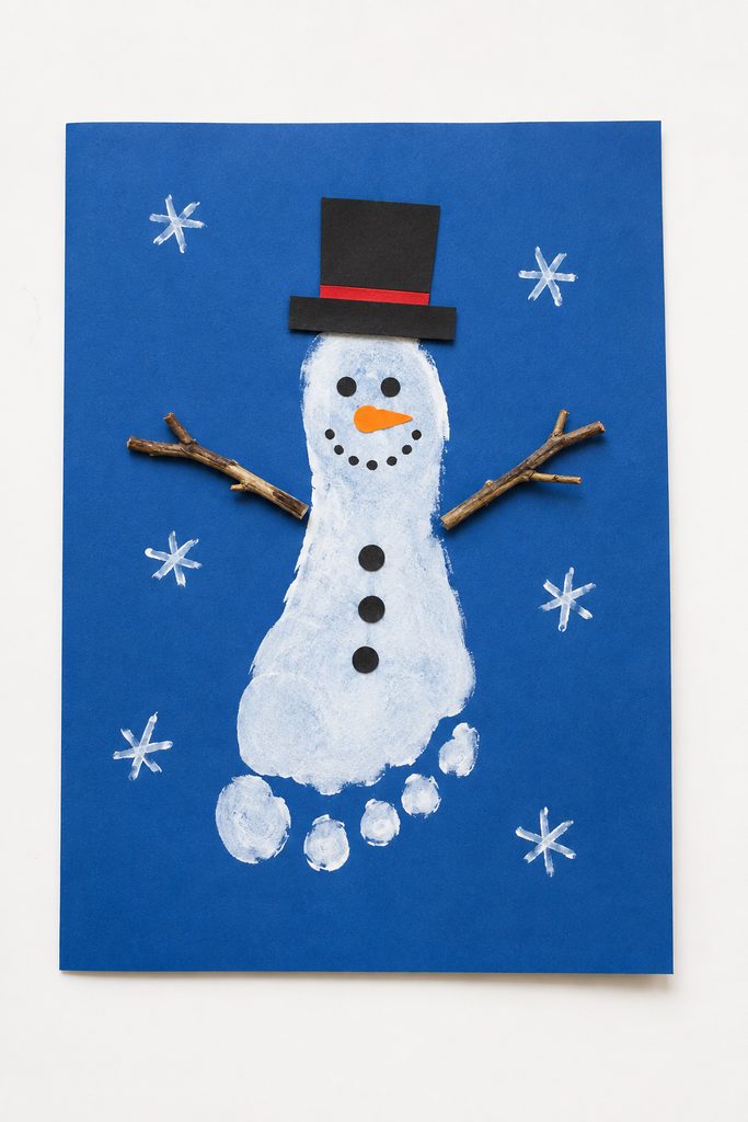 a snowman footprint craft made from a pale painted footprint on blue paper with a black hat, dot eyes, an orange nose, buttons, and twig arms on a white table
