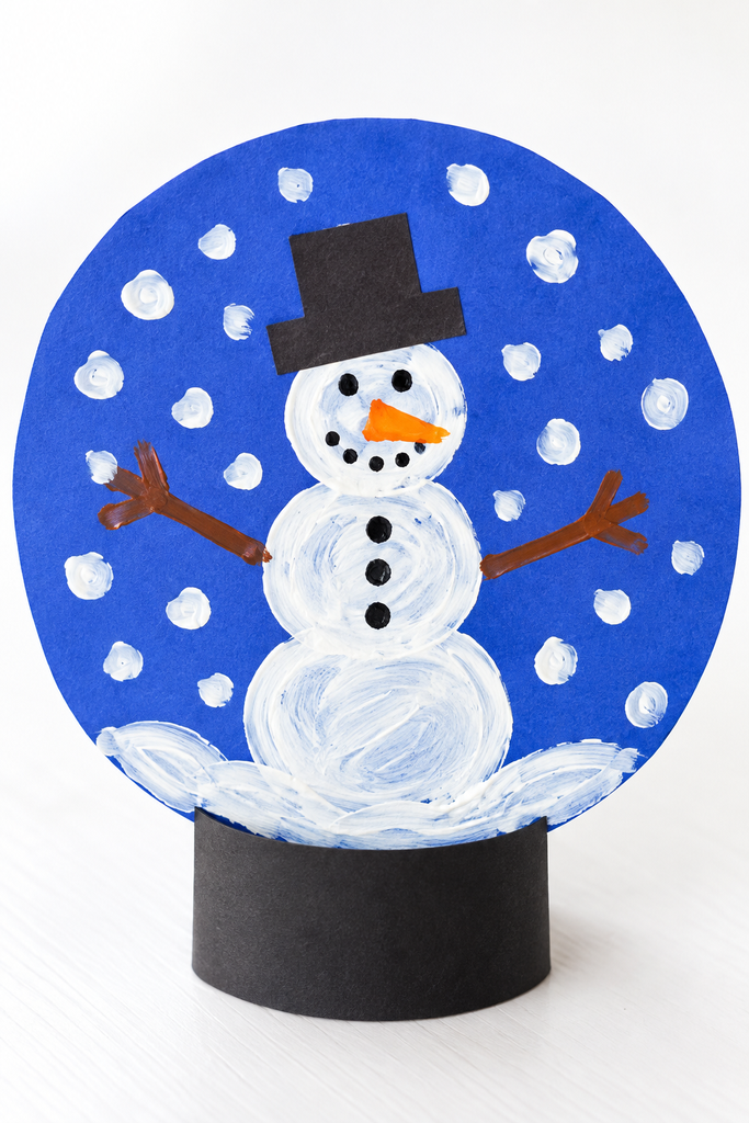 a paper snow globe craft with a little snowman inside on blue paper, white snow dots, and a black base on a white table