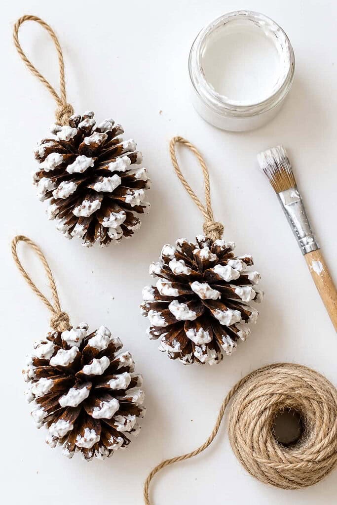 Frosted white-tipped pinecone ornaments with twine loops beside white paint, a brush, and twine on a white table.