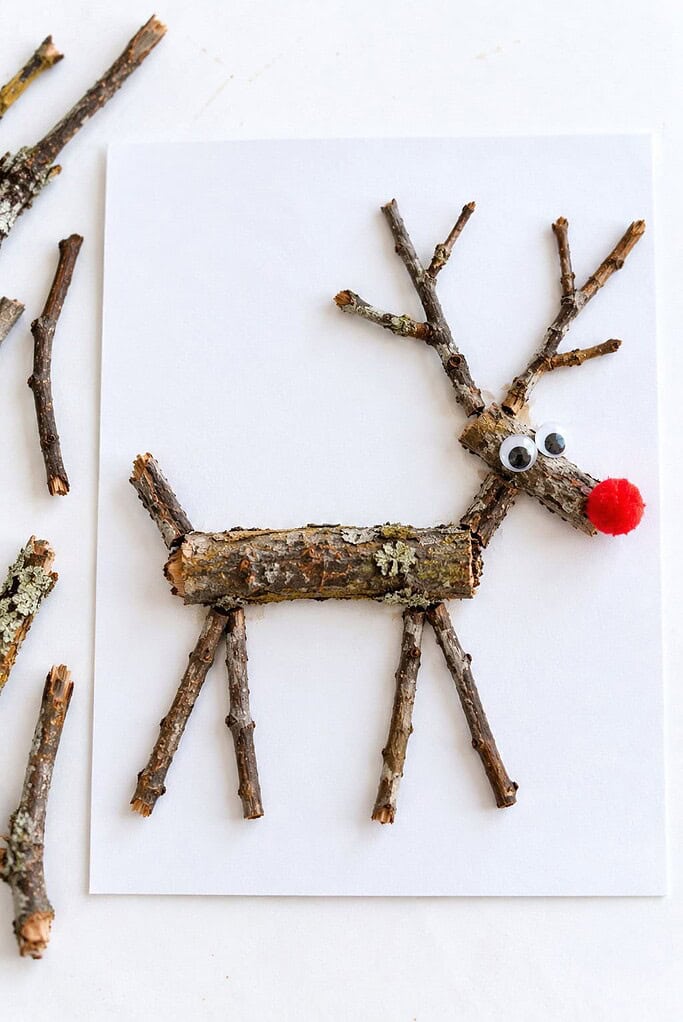 A reindeer made of real twigs glued on white cardstock with googly eyes and a red pom-pom nose, beside a few loose twigs.