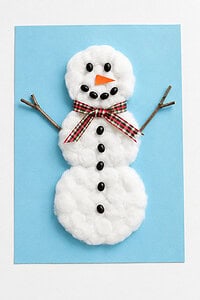 a handmade cotton-ball snowman craft with twig arms, a ribbon scarf, and a black bean face on light blue paper