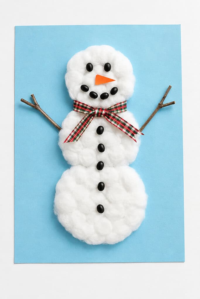 a handmade cotton-ball snowman craft with twig arms, a ribbon scarf, and a black bean face on light blue paper