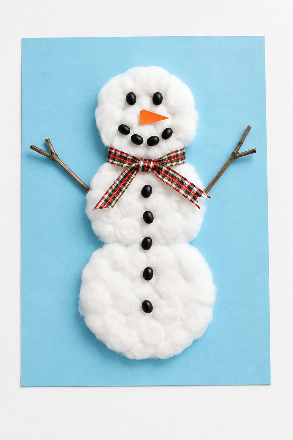 a handmade cotton-ball snowman craft with twig arms, a ribbon scarf, and a black bean face on light blue paper