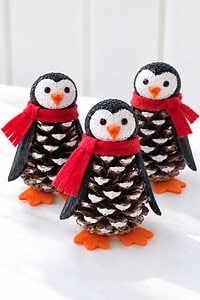 Three pinecone penguins with red scarves and orange feet grouped together on a bright white table.