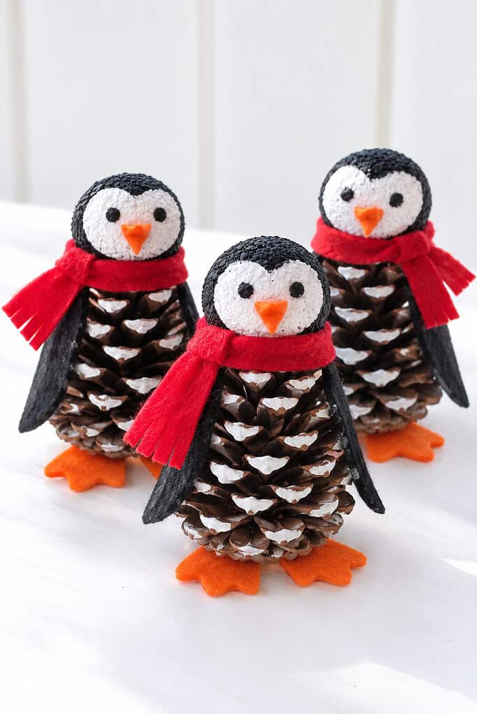 Three pinecone penguins with red scarves and orange feet grouped together on a bright white table.