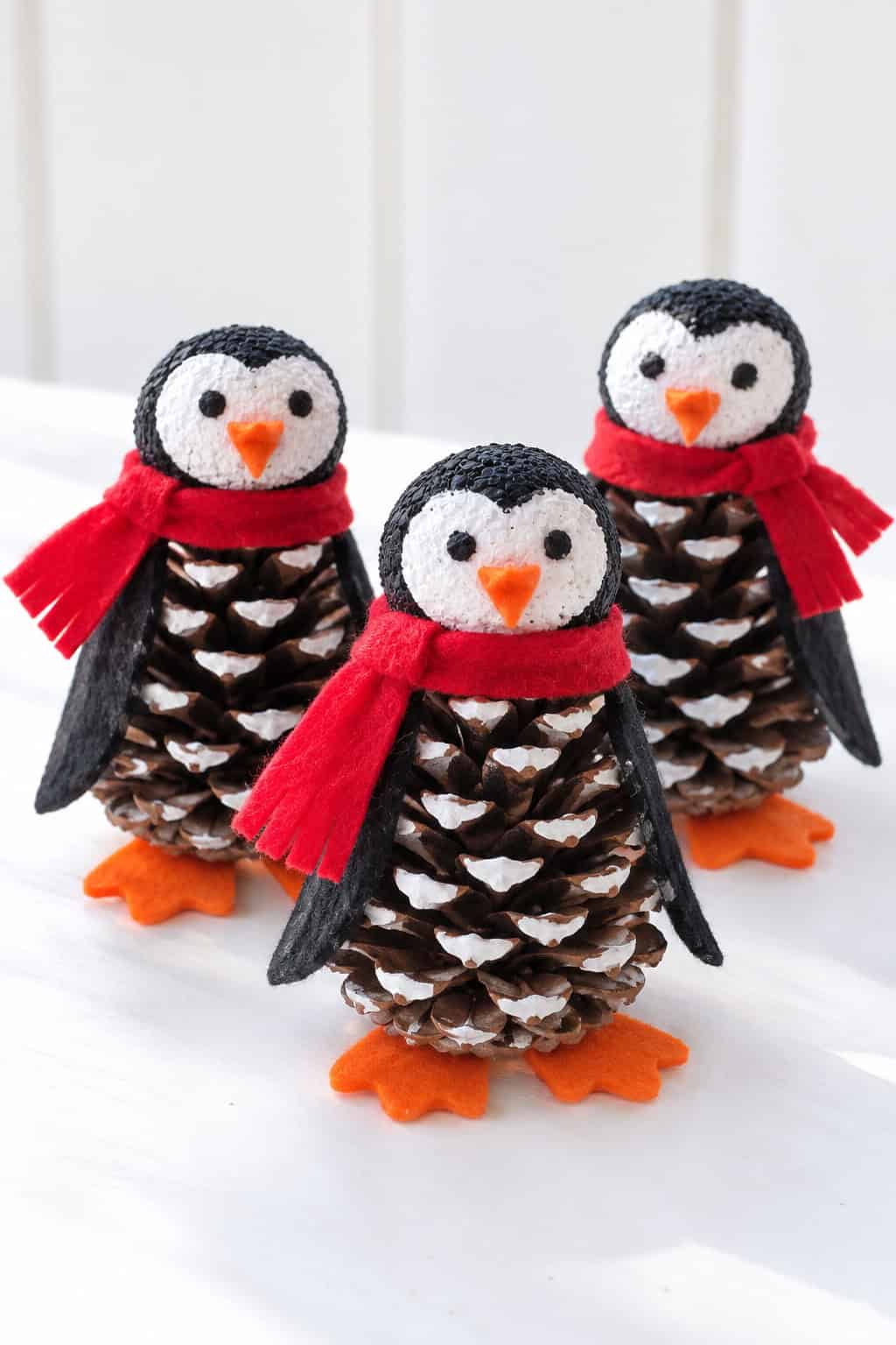 Three pinecone penguins with red scarves and orange feet grouped together on a bright white table.