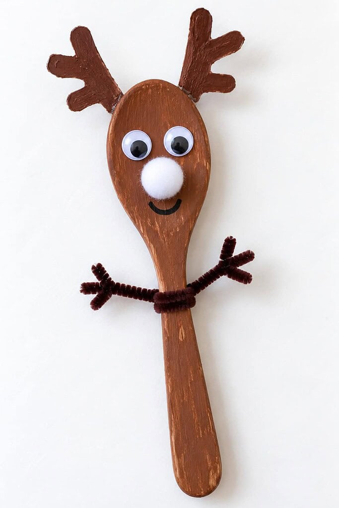 a wooden spoon reindeer craft painted brown with antlers, googly eyes, a white pom-pom nose, and pipe cleaner arms on a white table