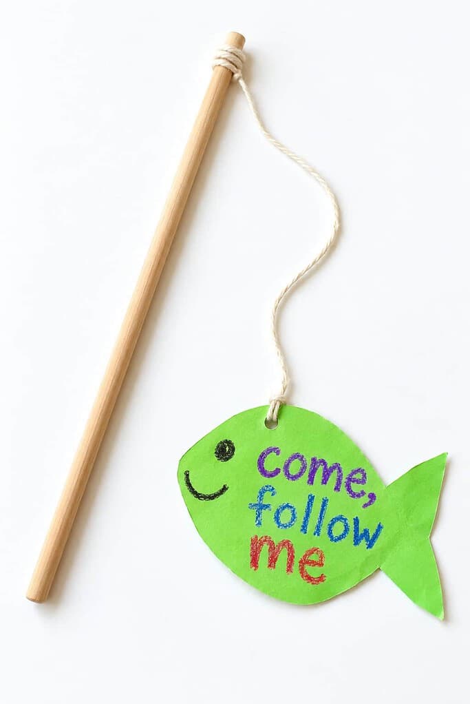 A wooden dowel fishing pole with a string and one bright green paper fish tied to the end, lettered come follow me.