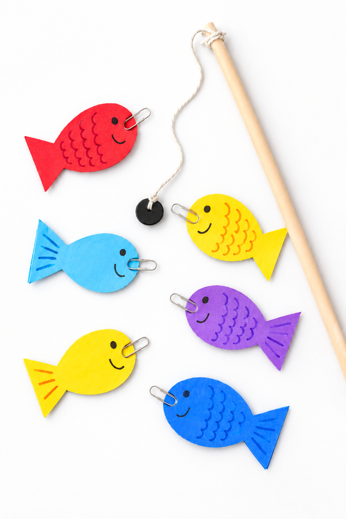 A dowel fishing rod with a magnet beside a scattered school of red, yellow, blue, and purple paper fish, each with a silver paperclip, on blue paper water.